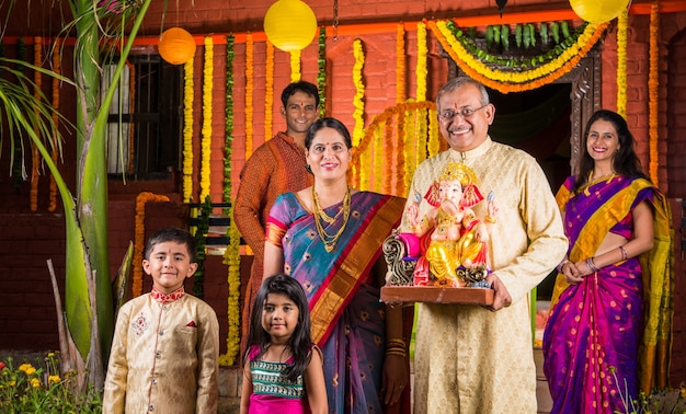 Joyeuse famille indienne célébrant le festival de Ganesh ou Chaturthi Accueillant ou jouant Pooja et mangeant des bonbons en tenue traditionnelle à la maison décorée de fleurs de souci