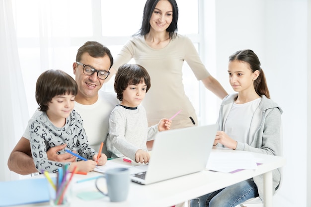 Joyeuse famille latine avec des enfants qui passent du temps ensemble à la maison. Père regardant des enfants jouer, dessinant tout en travaillant à domicile, utilisant un ordinateur portable. Technologie, concept de famille. Mise au point sélective