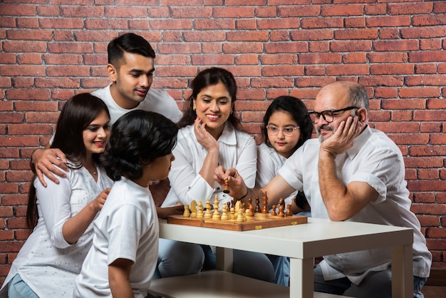 Joyeuse famille multigénérationnelle asiatique indienne de six personnes jouant aux échecs ou Shatranj qui est un jeu de société populaire, portant des vêtements blancs contre un mur de briques rouges