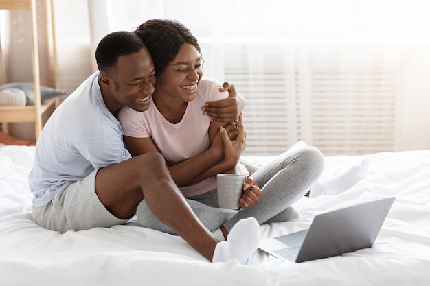 Joyeuse famille noire assise devant un ordinateur portable sur le lit et s'embrassant, buvant du thé et regardant un film comique, passant une journée de farniente ensemble. Joyeux couple afro-américain amoureux utilisant un ordinateur portable, espace vide