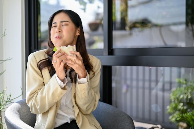 Joyeuse femme d'affaires asiatique heureuse et affamée appréciant son délicieux sandwich au café