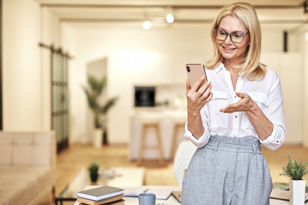 Joyeuse femme d'affaires mature dans des verres ayant un appel vidéo en ligne à l'aide d'un smartphone en position debout