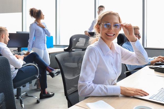 Joyeuse femme d'affaires en tenues de soirée travaillant sur un ordinateur portable dans le bureau moderne avec des collègues en arrière-plan