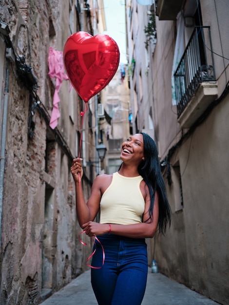 Joyeuse femme afro-américaine souriante tout en tenant un ballon coeur rouge à l'extérieur dans la rue. Concept de la Saint-Valentin.