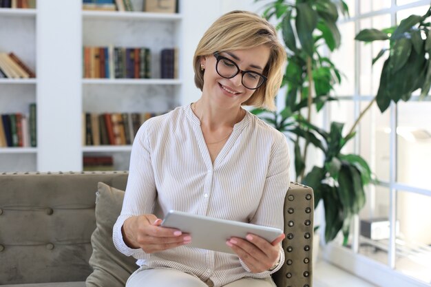 Joyeuse femme d'âge moyen assise sur un canapé, utilisant des applications pour tablettes informatiques, regardant l'écran, lisant de bonnes nouvelles sur les réseaux sociaux, faisant des achats ou discutant en ligne.