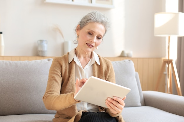 Joyeuse femme d'âge moyen assise sur un canapé, utilisant des applications pour tablettes informatiques, regardant l'écran, lisant de bonnes nouvelles sur les réseaux sociaux, faisant des achats ou discutant en ligne.