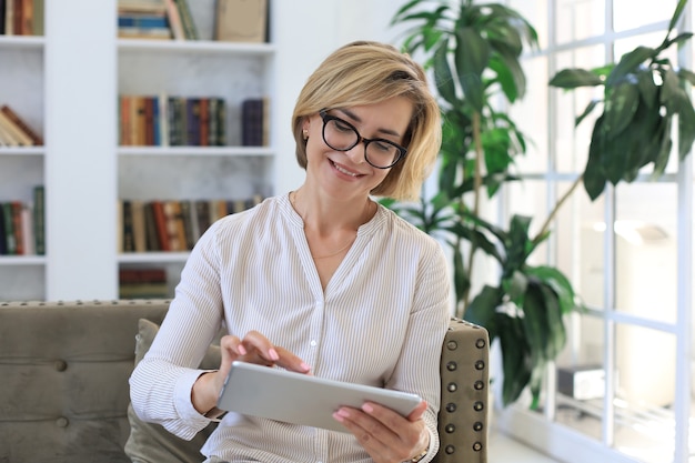 Joyeuse femme d'âge moyen assise sur un canapé, utilisant des applications pour tablettes informatiques, regardant l'écran, lisant de bonnes nouvelles sur les réseaux sociaux, faisant des achats ou discutant en ligne.