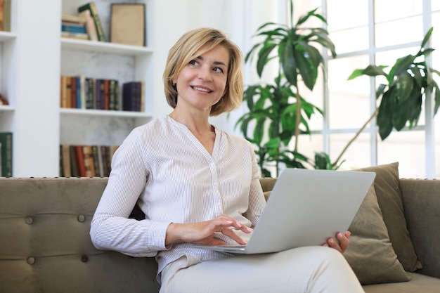 Joyeuse femme d'âge moyen utilisant un ordinateur portable assis sur un canapé à la maison.