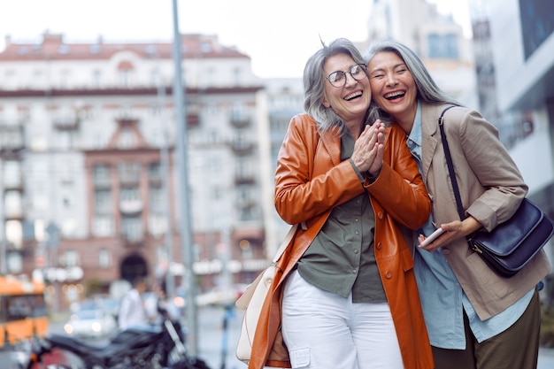 Joyeuse femme âgée et compagnon asiatique tenant un support de smartphone dans une rue de la ville moderne