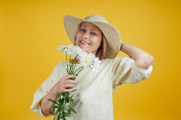 Joyeuse femme âgée dans un chapeau de paille avec des marguerites dans ses mains souriant sur un fond jaune vif. Concept de vente d'été, vacances d'été