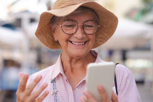 Joyeuse femme âgée séduisante marchant à l'extérieur dans la rue de la ville, parlant sur un téléphone portable, heureuse dame âgée avec chapeau d'été et lunettes, appel vidéo avec téléphone portable