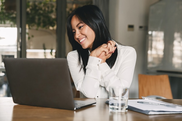 Joyeuse femme asiatique des années 20 portant une chemise blanche souriant et faisant des gestes de côté, tout en regardant l'écran de l'ordinateur portable au bureau
