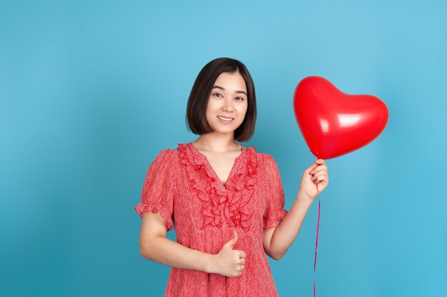 Joyeuse femme asiatique dans une robe rouge tient un ballon volant en forme de coeur rouge et donne un coup de pouce