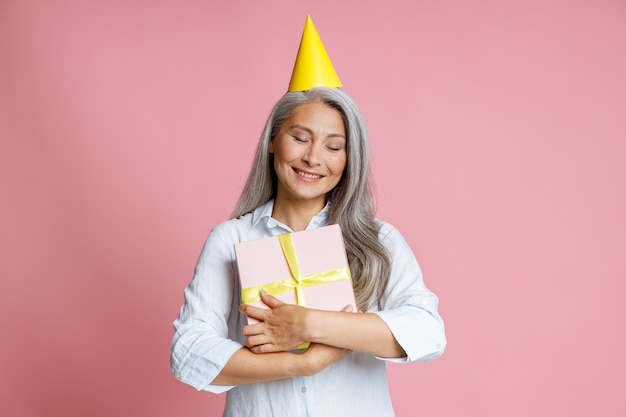 Joyeuse femme asiatique mature avec des cheveux gris lâches embrasse une boîte-cadeau qui pose en studio