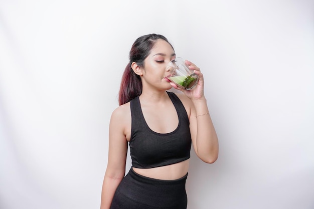 Joyeuse femme asiatique sportive portant des vêtements de sport boit un verre de délicieux smoothie vert isolé sur fond blanc