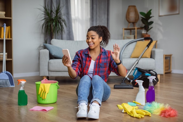 Photo joyeuse femme au foyer afro-américaine du millénaire agitant la main au smartphone assis sur le sol