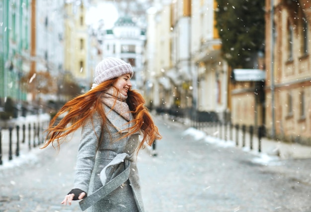 Joyeuse femme aux cheveux longs au gingembre porte un manteau élégant marchant dans la rue pendant les vacances de Noël avec de la neige. Espace pour le texte