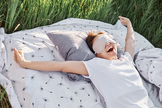 Joyeuse femme béante portant un pli endormi et un t-shirt décontracté blanc étirant les bras au réveil le matin au lit dans un pré vert, se relaxant et dormant en plein air dans le champ.