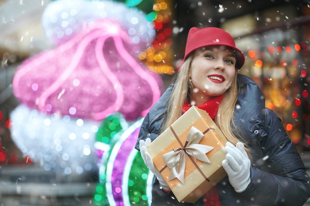 Joyeuse femme blonde avec une boîte-cadeau au marché de Noël de la rue pendant les chutes de neige. Espace libre