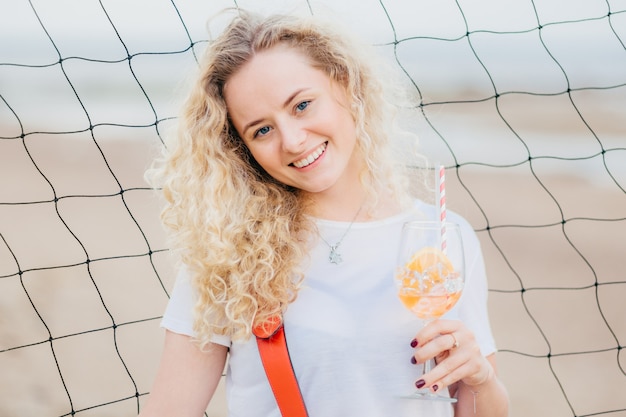 Joyeuse femme blonde heureuse vêtue de vêtements décontractés, détient un cocktail d'orange frais, sourit positivement