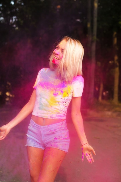 Joyeuse femme blonde en t-shirt blanc et short en jean sur le festival Holi dans le parc