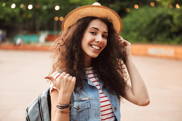 Joyeuse femme brune aux cheveux bruns bouclés, souriant et pointant le doigt de côté tout en marchant dans un endroit hipster ou un parc moderne