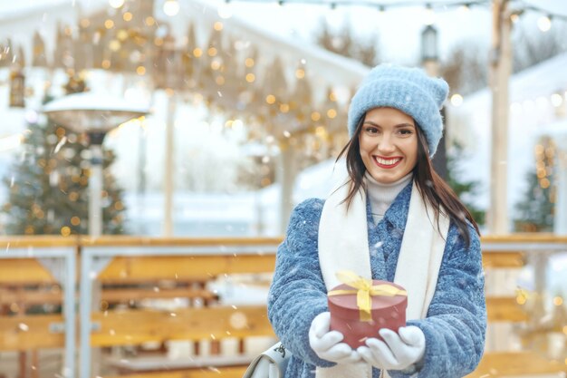 Joyeuse femme brune en manteau d'hiver tenant une boîte-cadeau à la foire de noël pendant les chutes de neige. Espace pour le texte