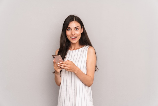 joyeuse femme brune portant une robe souriante tout en tenant un smartphone moderne isolé sur un mur gris