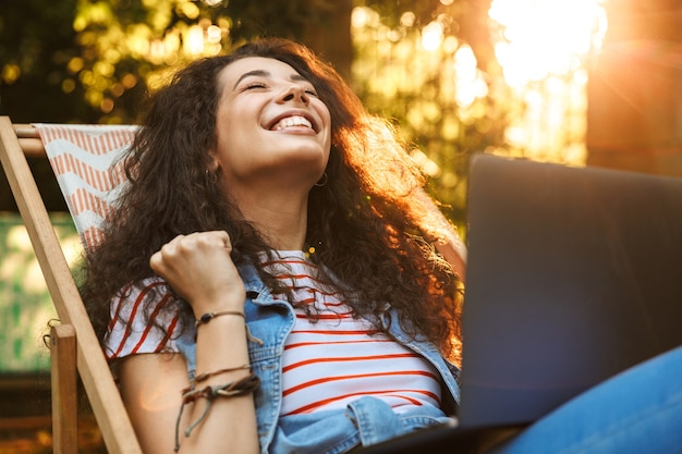 Joyeuse femme brune, serrant le poing de joie alors qu'il était assis dans une chaise longue pendant le repos dans le parc par une journée ensoleillée et à l'aide d'un ordinateur portable argenté avec surprise