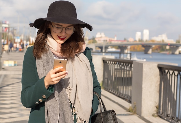 Joyeuse femme brune souriante à lunettes et chapeau vêtue de vêtements à la mode à l'aide d'un téléphone portable