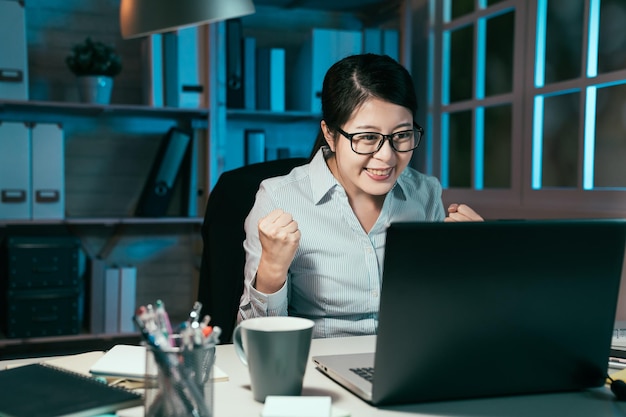 Joyeuse femme de bureau chinoise asiatique excitée assise à une table de travail et regardant l'écran d'un ordinateur portable. une employée fait un geste oui tout en terminant le travail à minuit sur le lieu de travail.