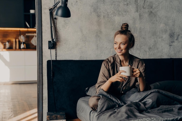 Joyeuse femme caucasienne souriante et heureuse en pyjama douillet en satin tenant une tasse de café