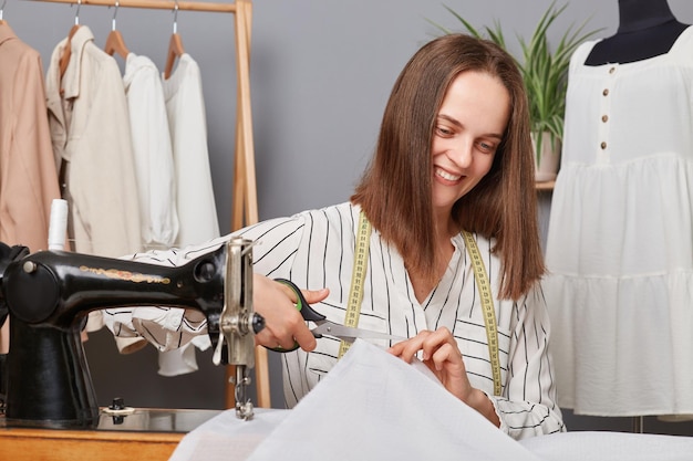 Joyeuse femme couturière coupant le textile dans le studio de couture lieu de travail de la couturière d'égout coupe les détails de la robe souriant joyeusement designer créant une nouvelle collection