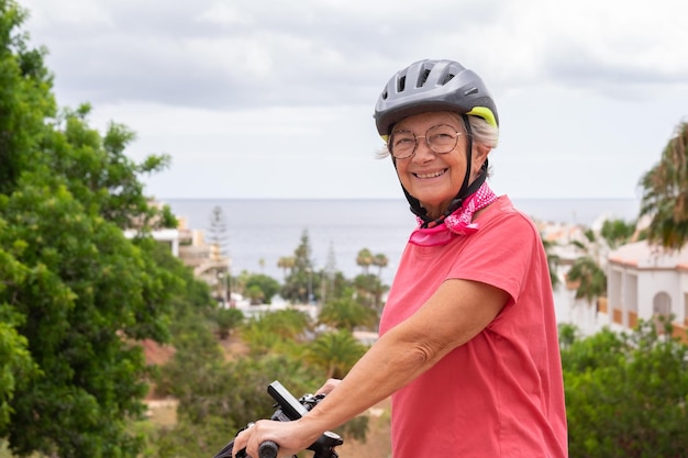 Joyeuse femme cycliste âgée en maillot rose et casque avec son vélo électrique sur la mer de la colline derrière profiter de la liberté et des vacances