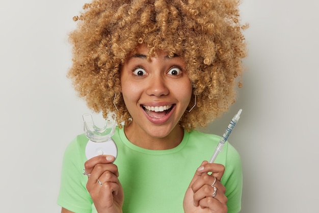 Joyeuse femme étonnée aux cheveux bouclés regarde avec joie la caméra tient un extenseur de bouche en plastique et une seringue avec du liquide vêtu d'un t-shirt vert décontracté isolé sur fond gris Hygiène dentaire