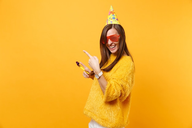 Joyeuse femme heureuse dans des lunettes orange drôles, chapeau d'anniversaire avec pipe en train de pointer les index de côté sur l'espace de copie, célébrant isolé sur fond jaune. Les gens émotions sincères, mode de vie.