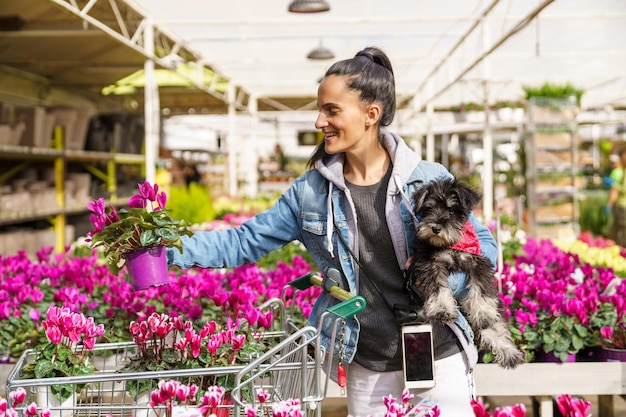 Joyeuse femme hispanique avec un chien noir mettant des fleurs de cyclamen en fleurs en pot dans votre panier tout en se tenant dans un magasin de fleurs léger