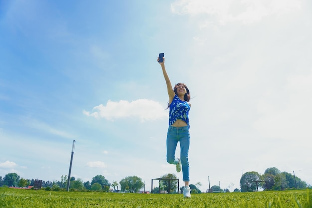 Joyeuse femme hispanique tenant un téléphone en l'air et courant dans un champ par une journée ensoleillée