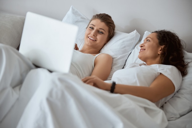 Joyeuse femme internationale allongée dans le lit et écoutant son heureux partenaire