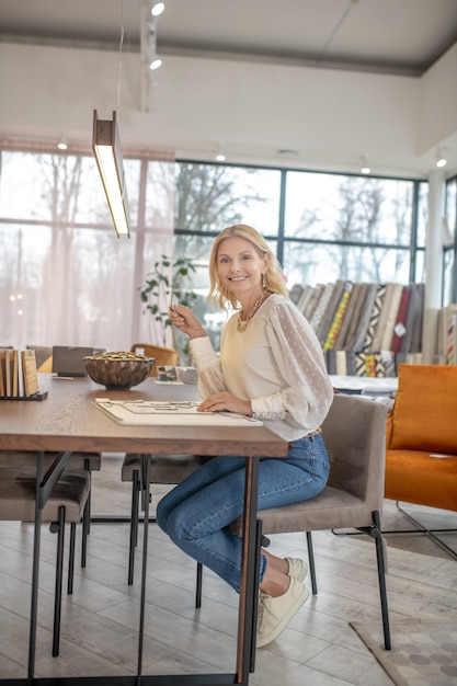 Joyeuse femme en jeans assis à une table dans une grande pièce lumineuse d'un magasin de meubles, souriant.