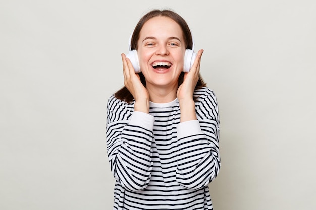Joyeuse femme joyeuse aux cheveux bruns portant une chemise rayée debout isolée sur fond gris répertoriant la musique préférée sur les écouteurs