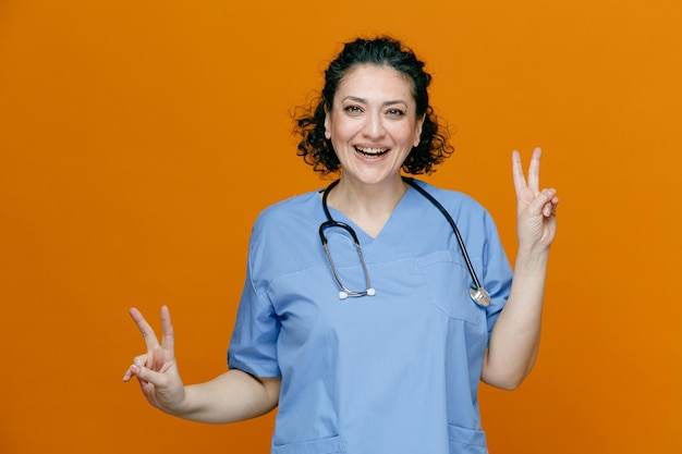 Joyeuse femme médecin d'âge moyen portant un uniforme et un stéthoscope autour du cou regardant la caméra montrant un signe de paix avec les deux mains isolées sur fond orange