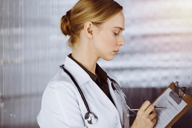 Joyeuse femme médecin souriante utilisant un presse-papiers dans une clinique ensoleillée. Portrait d'une femme médecin sympathique au travail. Notion de médecine.