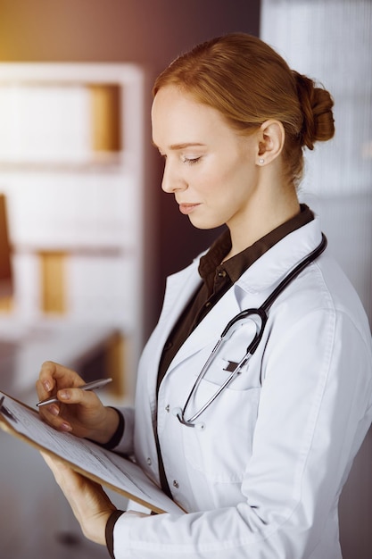 Joyeuse femme médecin souriante utilisant un presse-papiers dans une clinique ensoleillée. Portrait d'une femme médecin sympathique au travail. Notion de médecine.