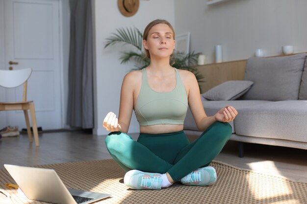 Joyeuse femme mince en survêtement assise sur le sol pratiquant le yoga en posture de lotus en gardant son calme à la maison.