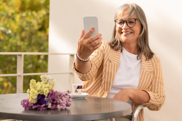 Joyeuse femme mûre européenne s'asseoir dans un café avec des fleurs boire du café prendre selfie sur smartphone