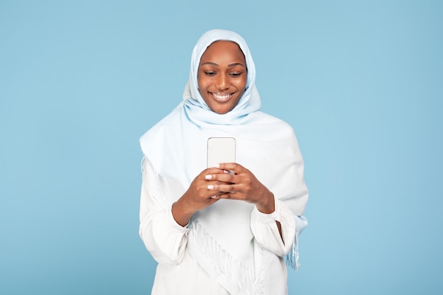 Joyeuse femme musulmane afro-américaine tenant un smartphone moderne regardant l'écran du gadget et souriant