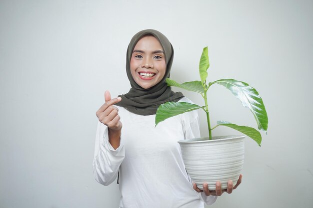Joyeuse femme musulmane asiatique en t-shirt et hijab tenant une plante et montrant un geste d'affection coréen je