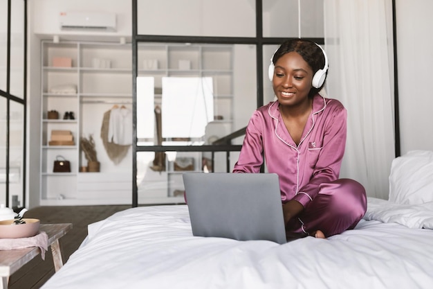 Joyeuse femme noire portant des écouteurs à l'aide d'un ordinateur assis dans la chambre