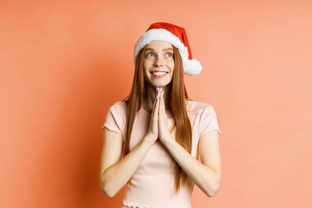 Joyeuse femme rousse aux taches de rousseur caucasienne en bonnet de Noel faisant un geste de prière, gardant les paumes ensemble, souriant largement, isolée sur fond de corail. Notion de langage corporel.